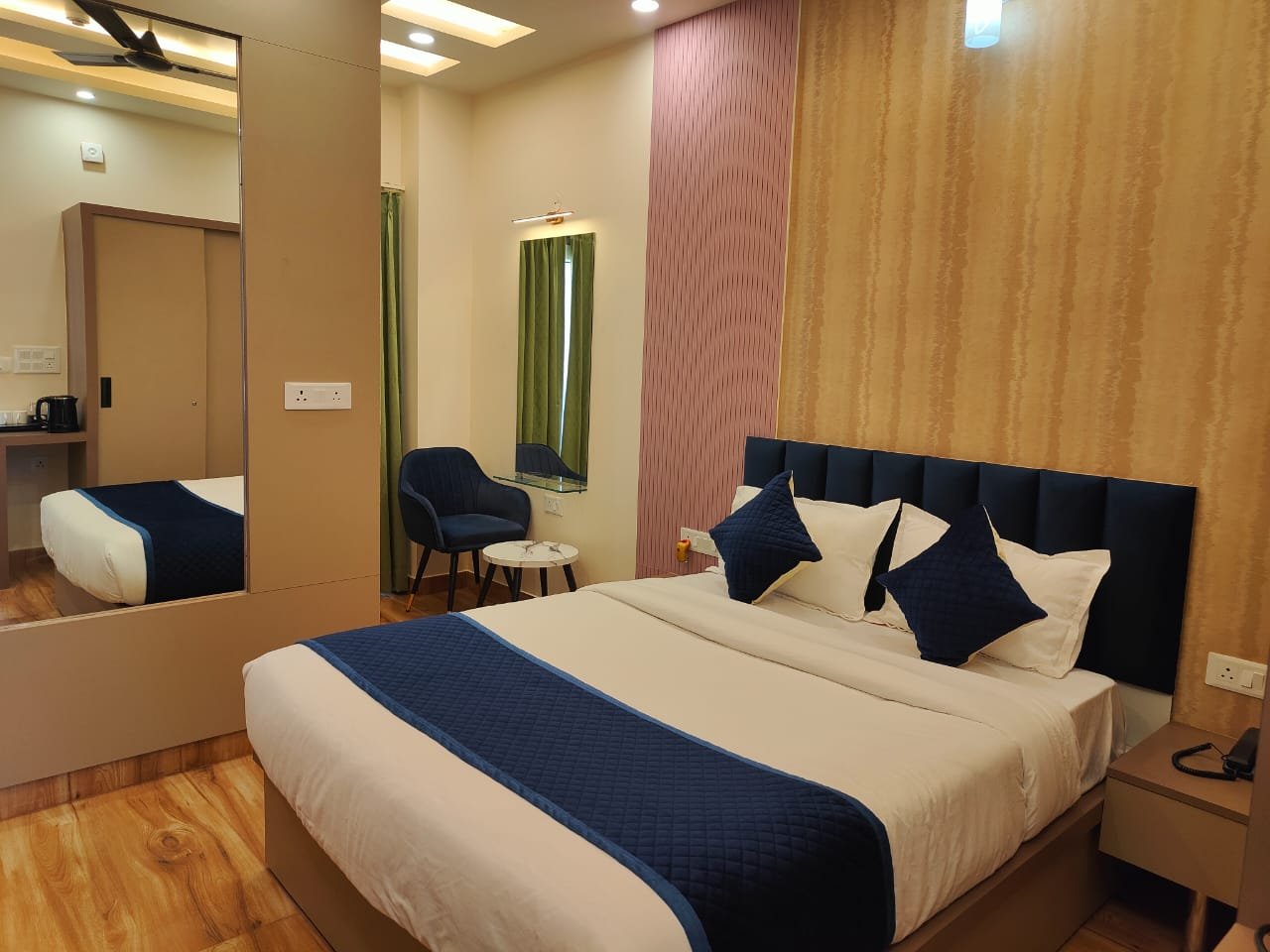 Luxurious hotel room at The Ummed Ahmedabad featuring elegant bed with colorful pillows, warm lighting, and modern furnishings in beige tones.